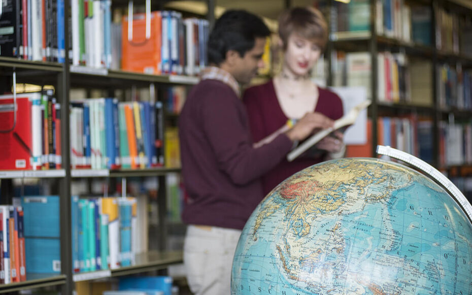 In der Hochschulbücherei stehen zwei Studierende im Hintergrund und lesen zusammen in einem Buch. Im Vordergrund steht ein Globus.