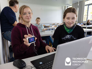 Zwei Mädchen stehen vor einem Laptop und eine hält Mikrocontroller in der Hand
