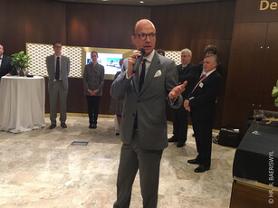 A man in a grey suit and glasses gives a speech with a microphone.