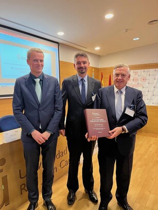 Three men in suits pose for a photo as one is presented with a book.