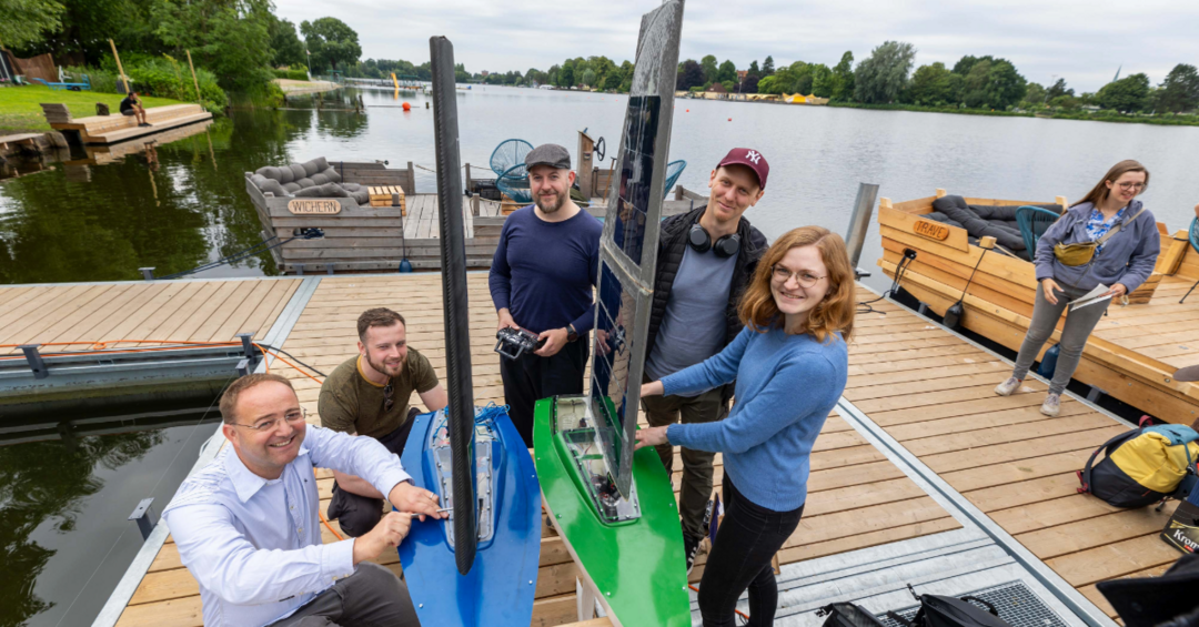 Studierendengruppe und Prof auf Steg an Fluss mit Solarsegelboot