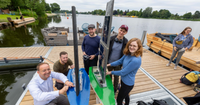 Studierendengruppe und Prof auf Steg an Fluss mit Solarsegelboot