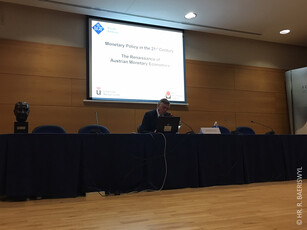 A man in a suit sits alone at a long table during a conference presentation.