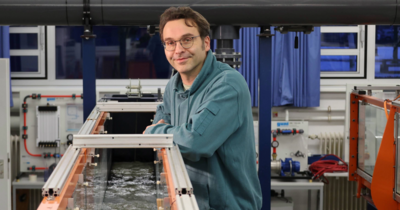 Ein Mann in einem türkisen Pullover lehnt an einem Wasserkanal, der in einem Wasserbaulabor steht. 