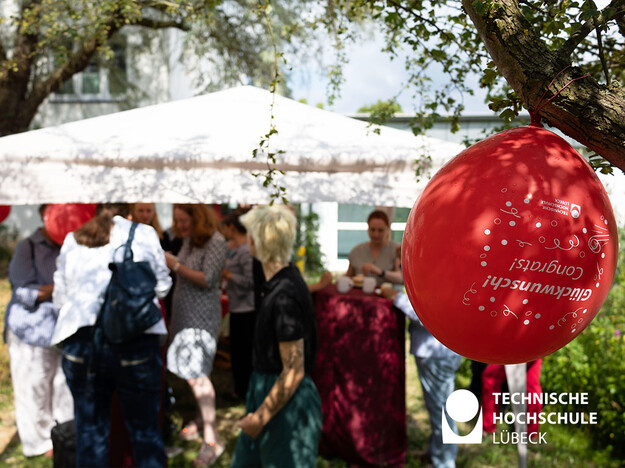 Bergfest beim Mentoringprogramm Im Fokus ein roter Luftballon und im Hintergrund mehrere Personen unter einem Pavillon