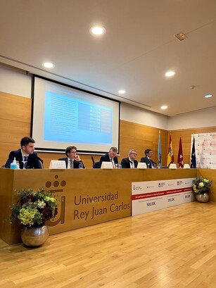 Five men are seated at a panel discussion in front of a screen with a chart.
