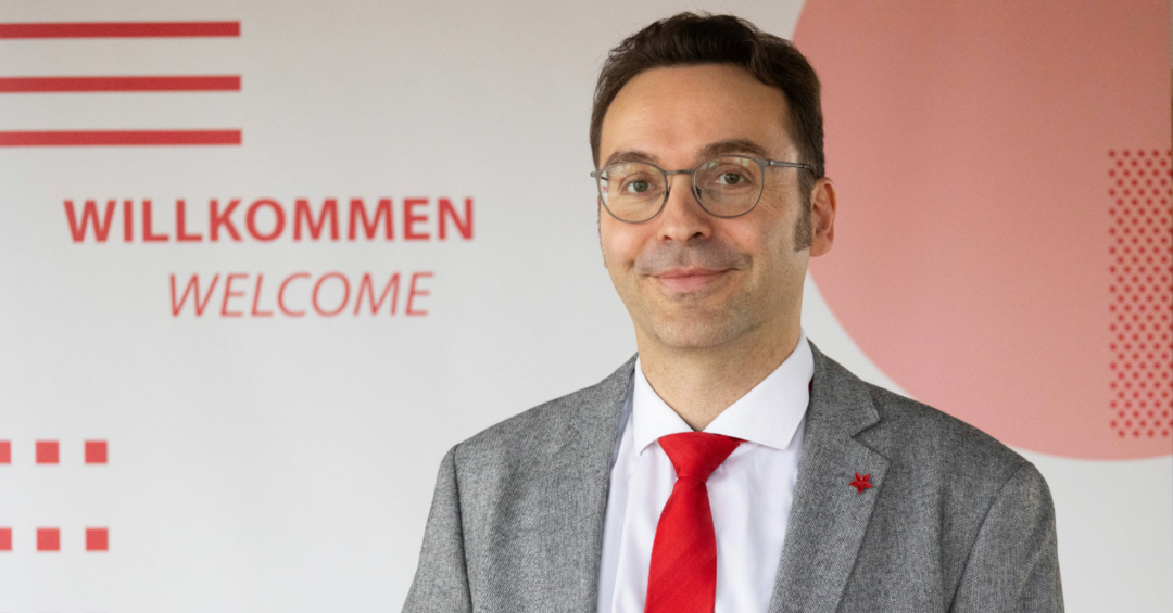 Ein Mann in grauem Anzug mit roter Krawatte und Brille lächelt in die Kamera