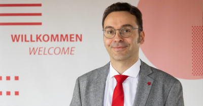 Ein Mann in grauem Anzug mit roter Krawatte und Brille lächelt in die Kamera