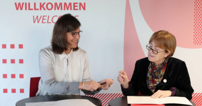 Zwei Frauen unterzeichnen lächelnd Dokumente an einem Tisch.