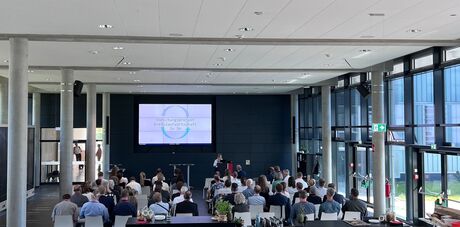 Panoramaaufnahme eines gut besuchten, modernen Konferenzraums mit Publikum, Bühne und großer Präsentationsleinwand.
