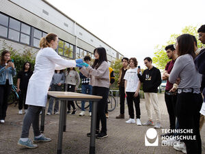 Eine Frau in weißem Kittel gibt einer Studentin zwei Kolben in die Hand, damit sie prüfen kann welche Temperatur diese haben