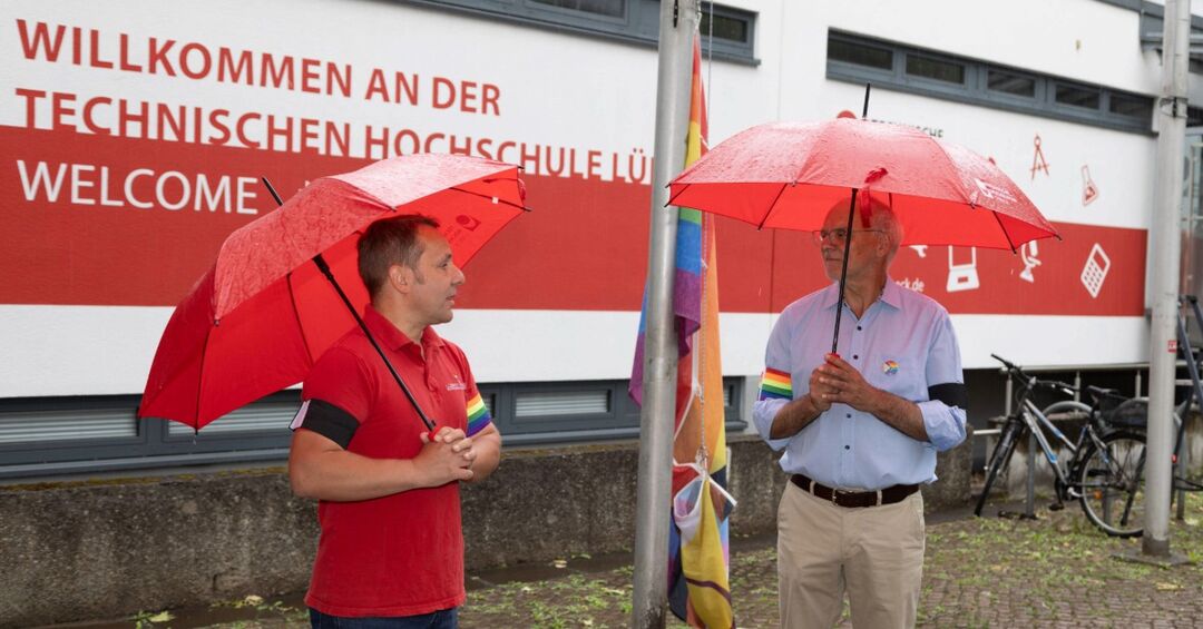 Christian Till und Jochen Abke stehen mit Regenschirmen neben der Pride-Flagge.
