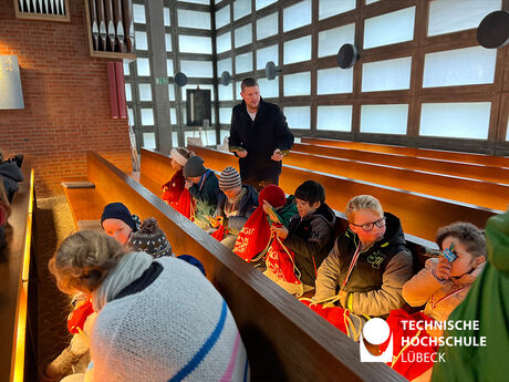 10 Kinder und 2 Erwachsene sitzen in zwei Reihen in einer Kirche und gucken in roten Beuteln hinein