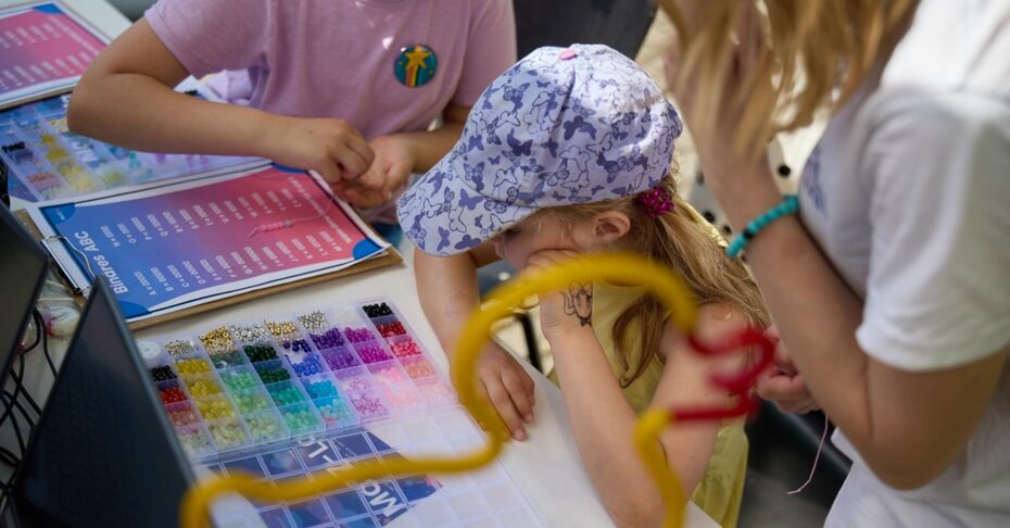 th_luebeck_juniorcampus_perlenketten Kinder sitzen an einem Tisch und fädeln bunte Perlen auf Schnüre. Neben ihnen liegt ein Arbeitsblatt mit einem binären Alphabet.