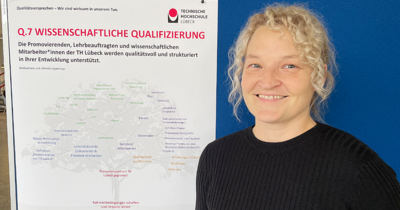 Eine Frau mit blonden, lockigen zurückgebundenen Haaren steht vor einer blauen Wand auf die ein Poster gepinnt ist