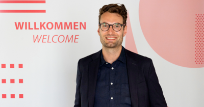 Ein Man vor einem rot weißen Hintergrund, der in die Kamera lächelt. Auf dem Hintergrund steht Willkommen/Welcome