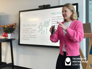 Eine Frau mit pinker Strickjacke steht vor einem Bildschirm und hält einen Vortrag