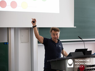 Experiment Rohre erzeugen Töne Ein Mann hält ein Plastikrohr in die Luft, er steht an einem Rednerpult und richtet sich an eine Menschenmenge