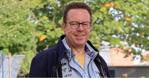 Ein Mann mit Brille steht draußen und lächelt in die Kamera 