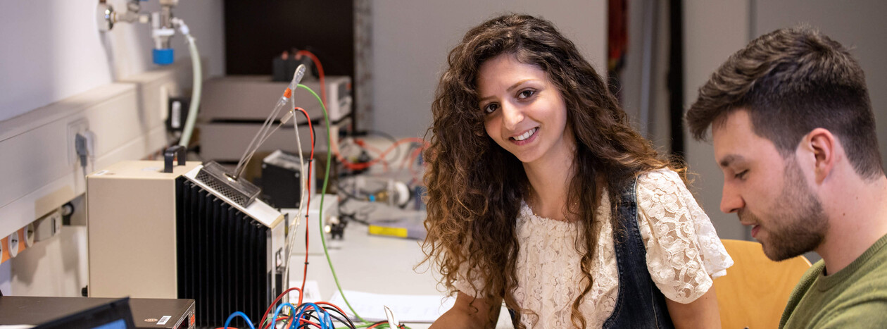 A female student smiles at the camera while working with a male student on an experiment involving electronic components and numerous wires in a technology lab.