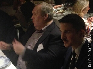 Two men in suits sit next to each other in conversation at a dinner.
