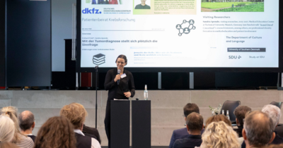 Eine Frau vor einer Leinwand, die zum Publikum spricht