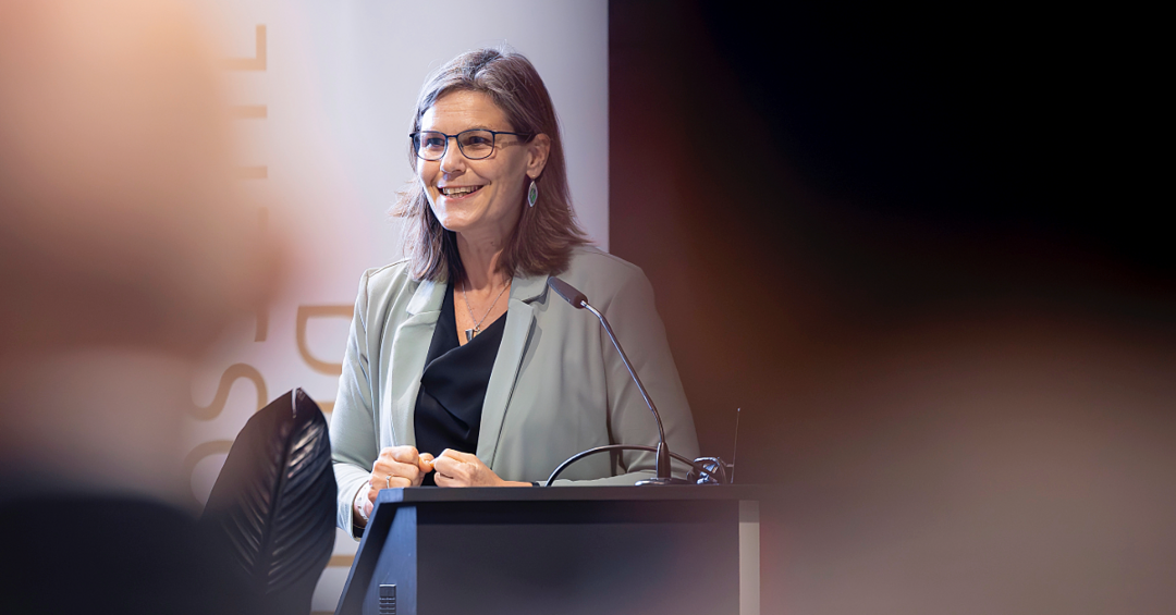 Eine Frau steht einem Pult, hat die Hände ineinandergefaltet und lächelt