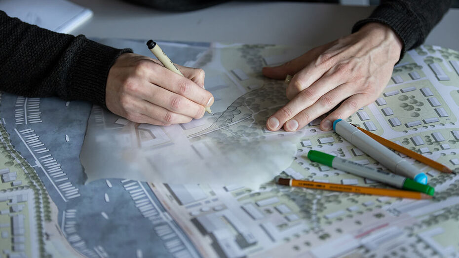 Auf einem Tisch ist ein Farbiger Stadtplan ausgerollt. Man sieht die Hände eines Studirenden und unterschiedliche Stifte liegen auf dem Plan. Die Hände zeichnen mit einer Schablone neue Elemente ein.