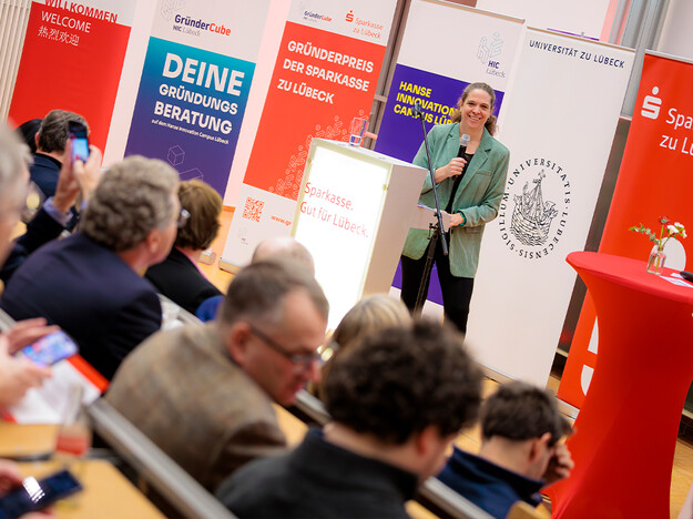 Moderation durch Geschäftsführerin der Hanse Innovation Campus GmbH Im Vordergrund Menschen, die im Hörsaal sitzen, im Hintergrund eine Frau, die moderiert.