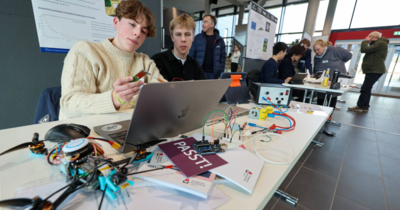 Hier sieht man zwei junge Männer, einen Laptop vor sich, eine Spielzeugdrohne im vorderen Bildausschnitt