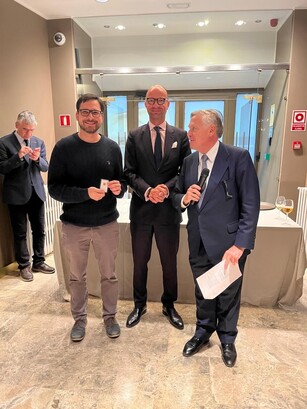 Three men pose; a man in a sweater holds an award, a presenter holds a mic.