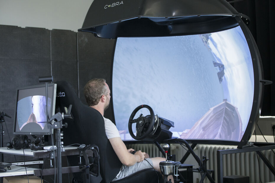 Ein großes Display mit einem Lenkrad davor. Ein Mensch sitzt vor dem Lenkrad und schaut gebannt auf die Simulation.