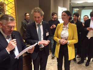 A presenter speaks while a woman in a yellow jacket and a man listen.