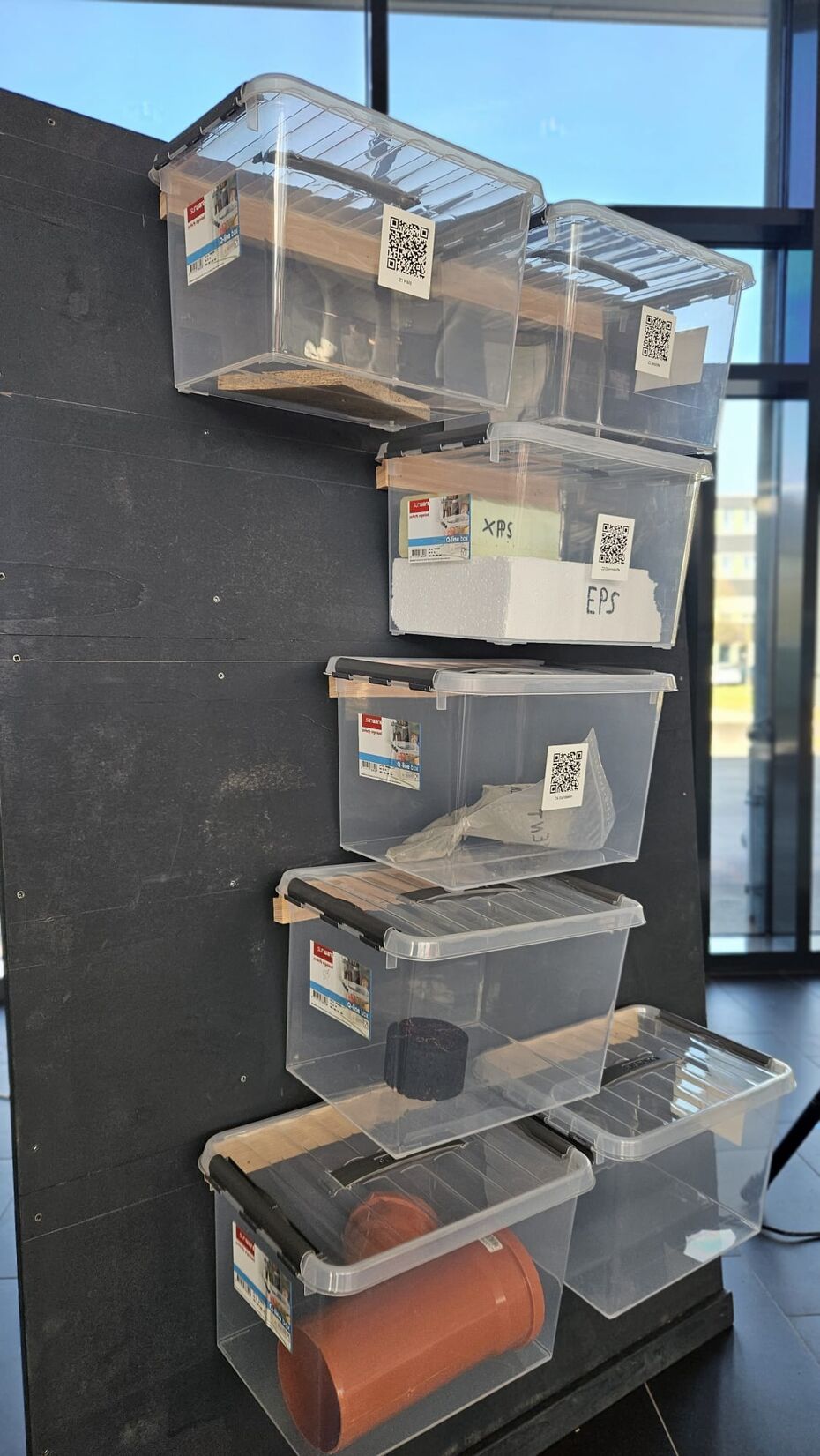 Transparente Boxen mit Bauprodukten und QR-Codes an einer Stellwand