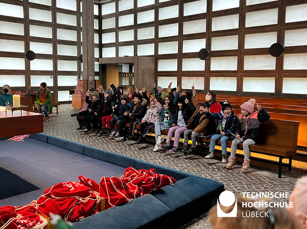 Viele Kinder sitzen auf einer Bank und gucken gespannt auf die roten Beutel mit den Geschenken