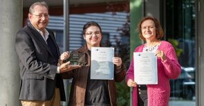 Drei Personen auf einem Bild, sie halten Urkunden vor sich und lächeln