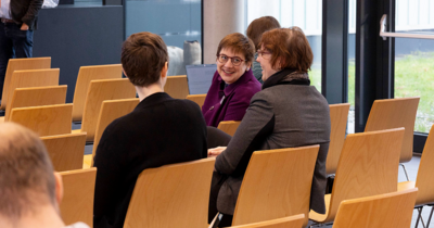 Drei Frauen sitzen in einer Stuhlreihe und lachen sich gegenseitig an