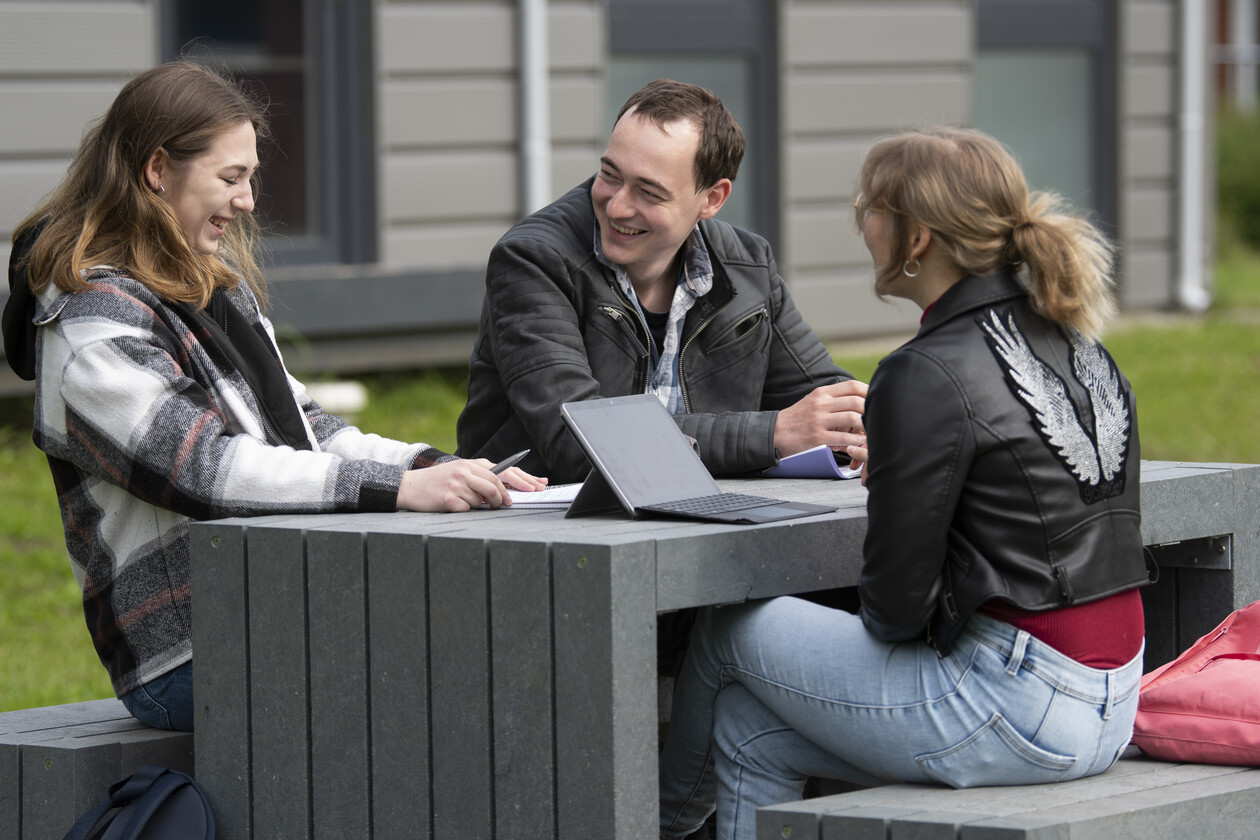 Drei Studierende sitzen draußen an einem Tisch, lachen und lernen zusammen.
