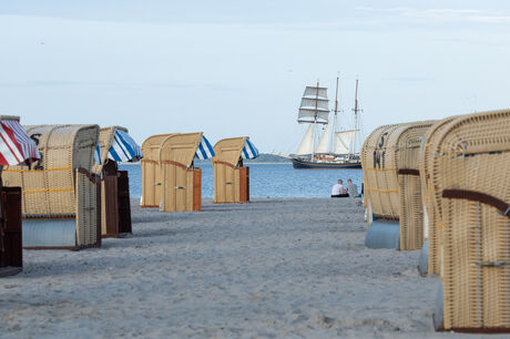 Strandkörbe im Sand mit Blick auf ein großes Segelschiff auf dem Meer.