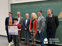 Bild zeigt Kind mit Check in der Hand und 6 weitere Personen hinter ihm die für das Foto posieren