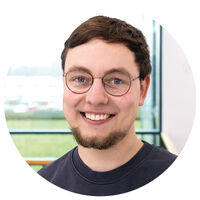 Junger Mann mit kurzen braunen Haaren, Bart und Brille lacht in die Kamera