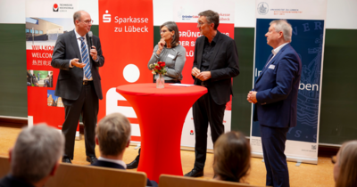 Vier Personen stehen auf einer Bühne an einem Stehtisch und unterhalten sich