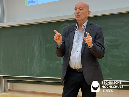 Studienabbruch Systemversagen oder selbstverschuldet Ein Mann steht vor einer grünen Tafel und gibt eine Vorlesung. Er hat eine Hand in die Luft gestreckt und spricht mit großer Leidenschaft. Der Mann trägt eine Jacke und einen Anzug.