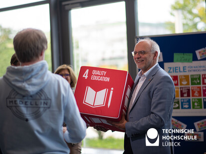 Nachhaltigkeit an der TH Lübeck Ein Mann in einem grauen Jackett hält einen großen, roten Würfel mit der Aufschrift "4 Quality Education" in den Händen. Er lächelt. Es gibt auch andere Menschen in der Szene, die sich um ihn versammelt haben.