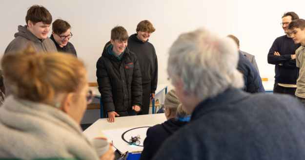 Hier sieht man im Hintergrund verschiedene Personen, die lachen und auf ein Blatt schauen, auf dem zwei Roboter Sumoringen