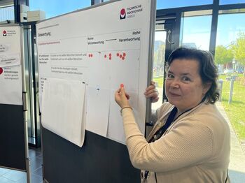 Frau klebt lächelnd einen roten Punkt auf ein Plakat zur KI-Bewertung.
