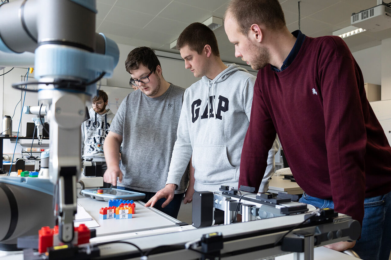 Drei Studierende beobachten die Messdaten eines Roboterarms auf einem Monitor. Der Roboterarm greift Bauklötze auf einem Labortisch