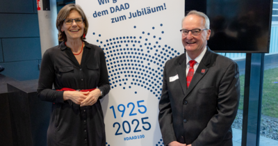 Eine Frau steht links neben einem Rollup auf dem DAAD steht, rechts daneben ein älterer Mann