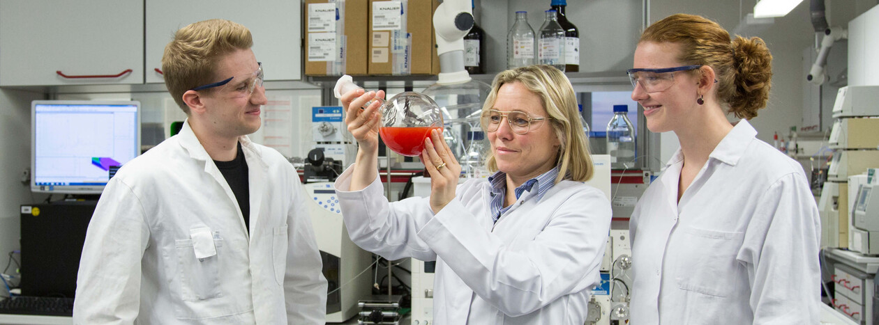 A female professor in a lab coat holds up a glass flask with a red liquid, discussing it with two students in a modern, well-equipped chemistry laboratory.