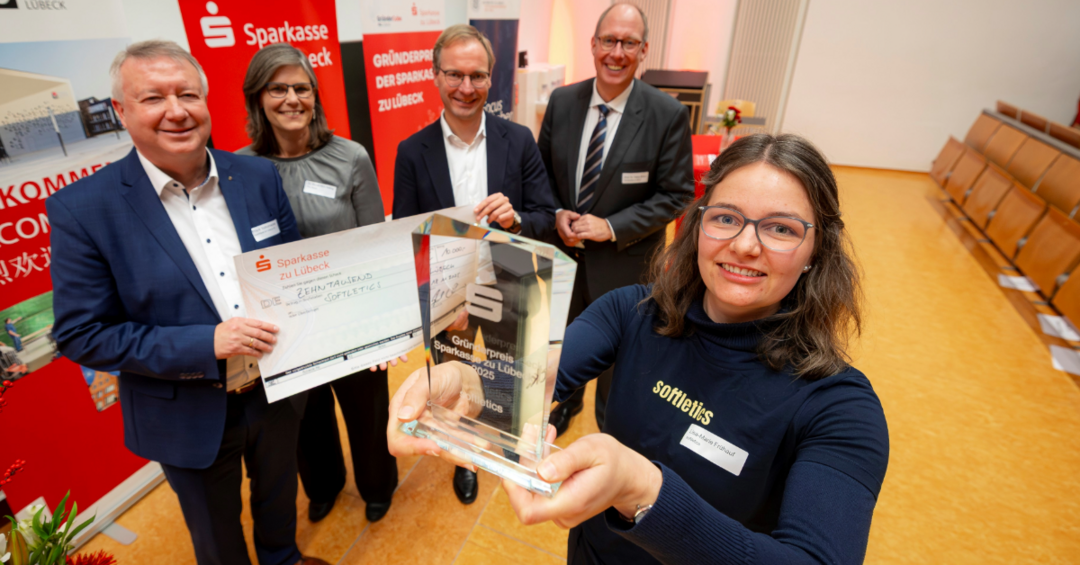 Eine Frau steht im Vordergrund und hält einen Preis aus Glas in die Kamera, hinter ihr vier Personen mit einem Scheck